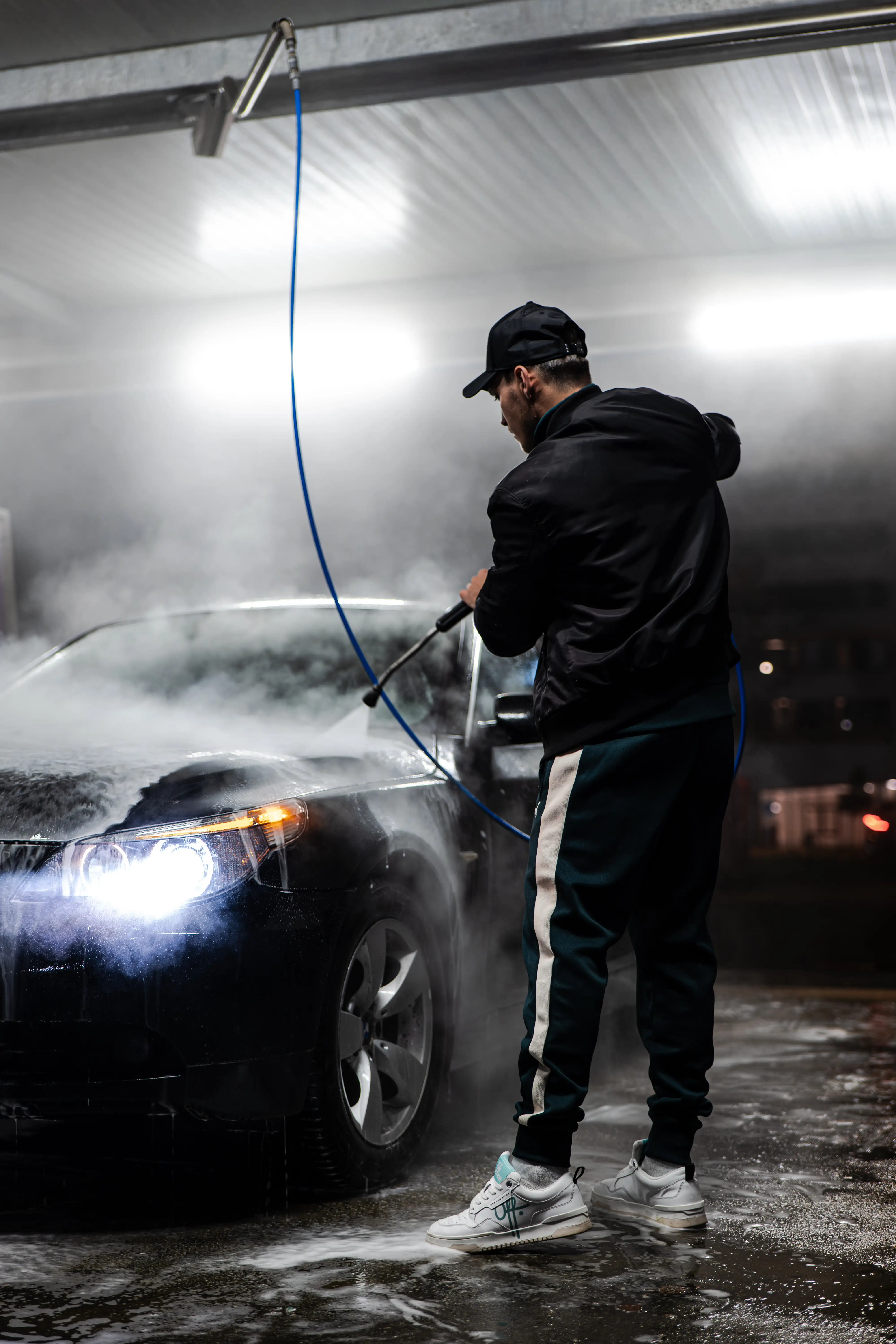Man pressure‑washing a black car at night, headlights glowing through steam, Zagreb.