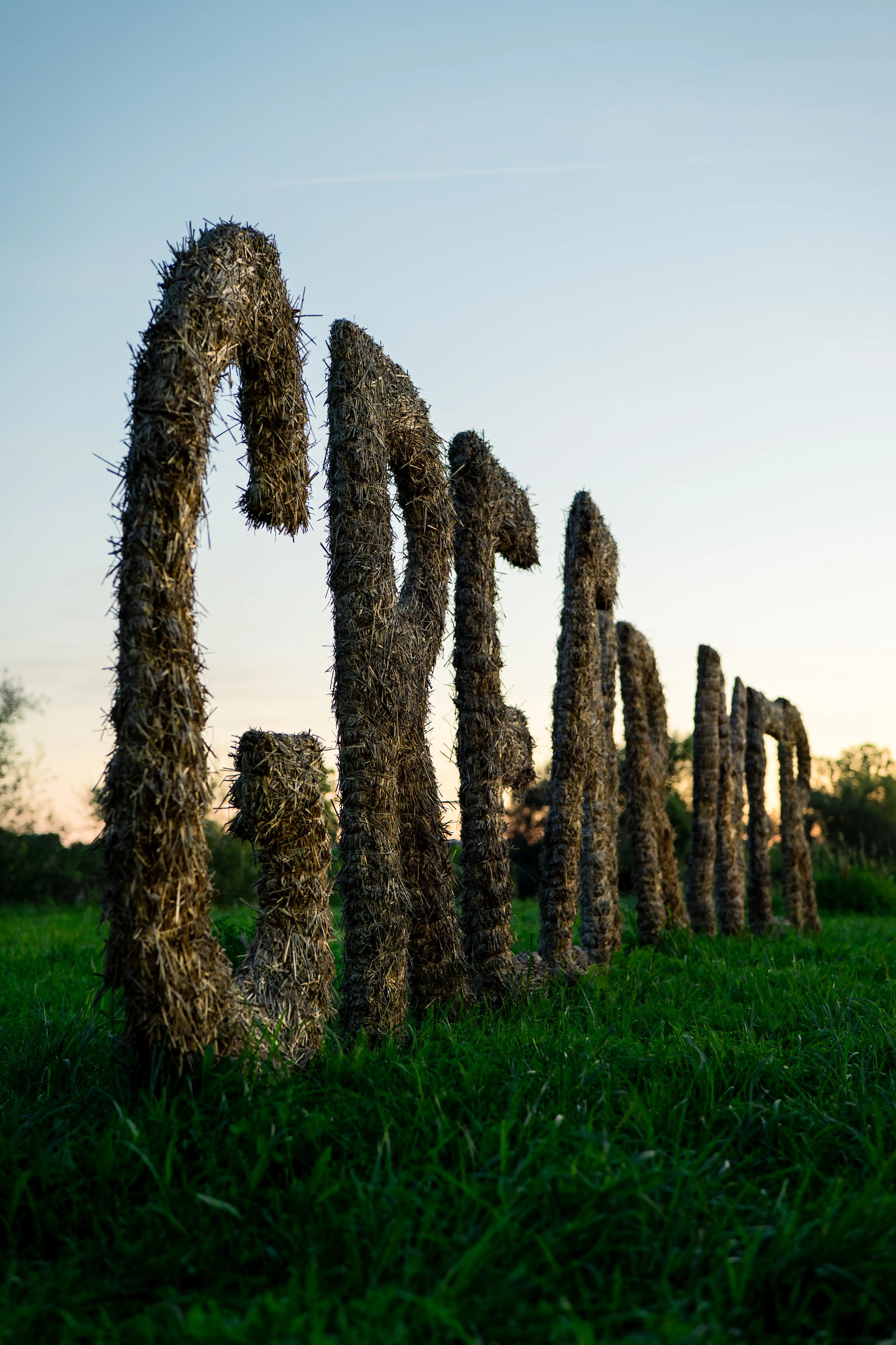 A low-angle, outdoor photograph of a large 'GREEN RIVER' sign, with letters sculpted from straw bales, standing in a vibrant green field against a clear sky at dusk for the Green River event.