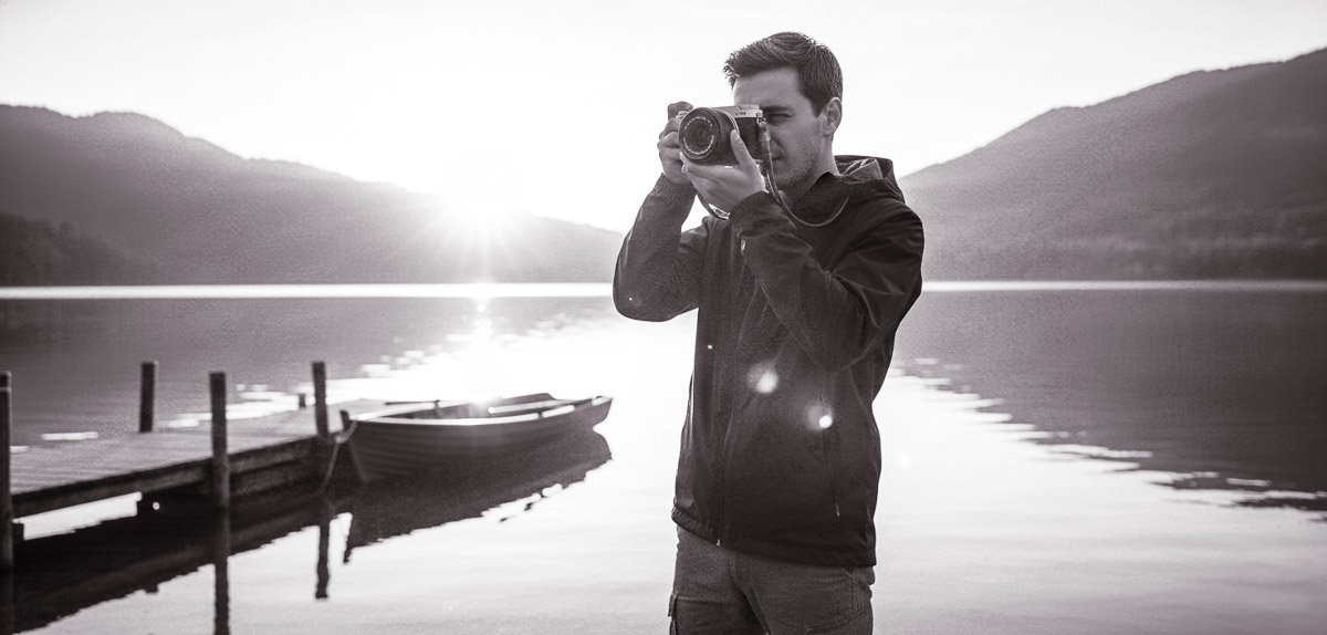 A low-angle cinematic shot of a person holding a silver and black Fujifilm X-M5 camera, standing by a serene lake and taking a picture of the misty mountains in the background during sunrise or dusk.