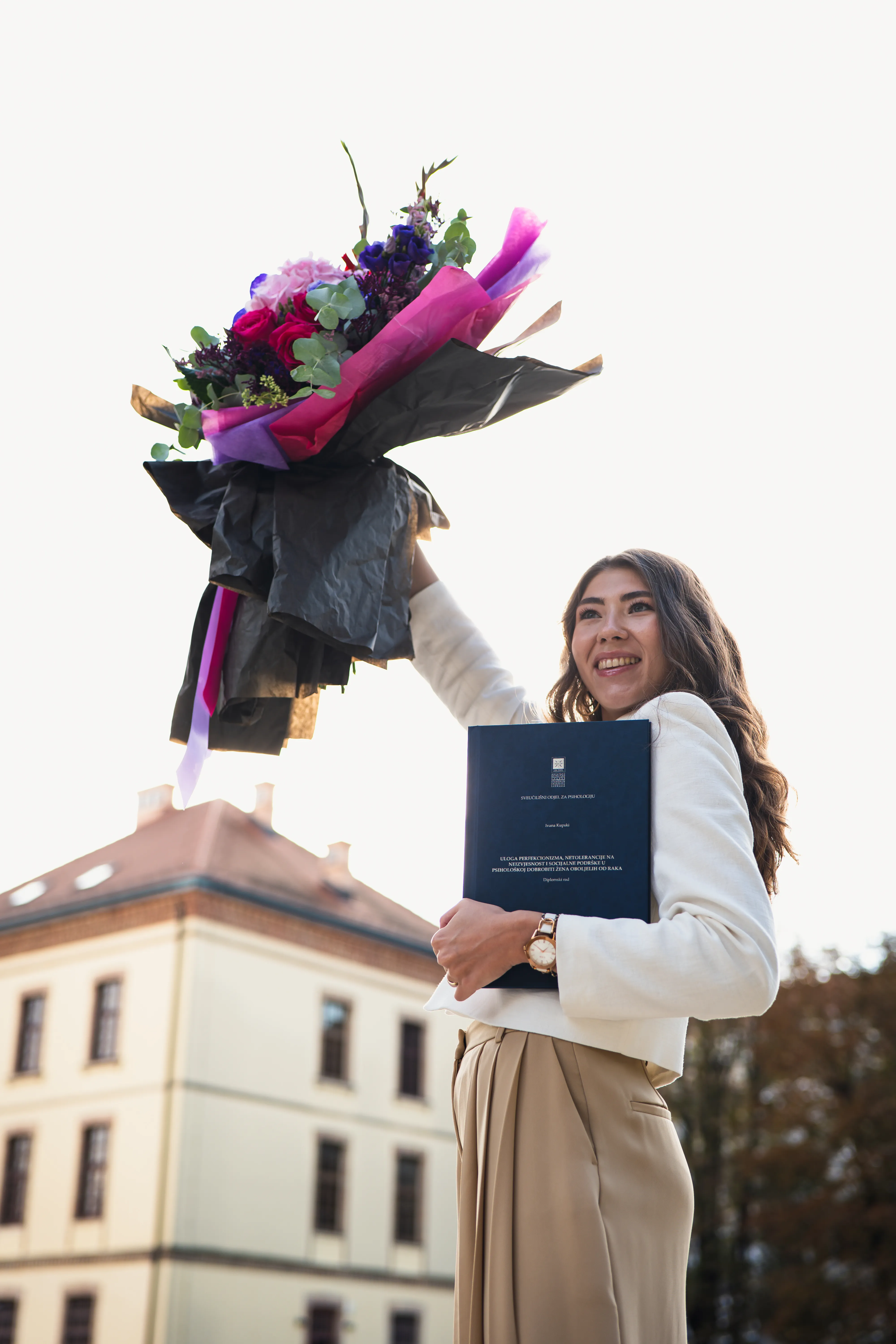 Joyful graduate raises flowers while clutching blue thesis folder during outdoor ceremony in Zagreb.