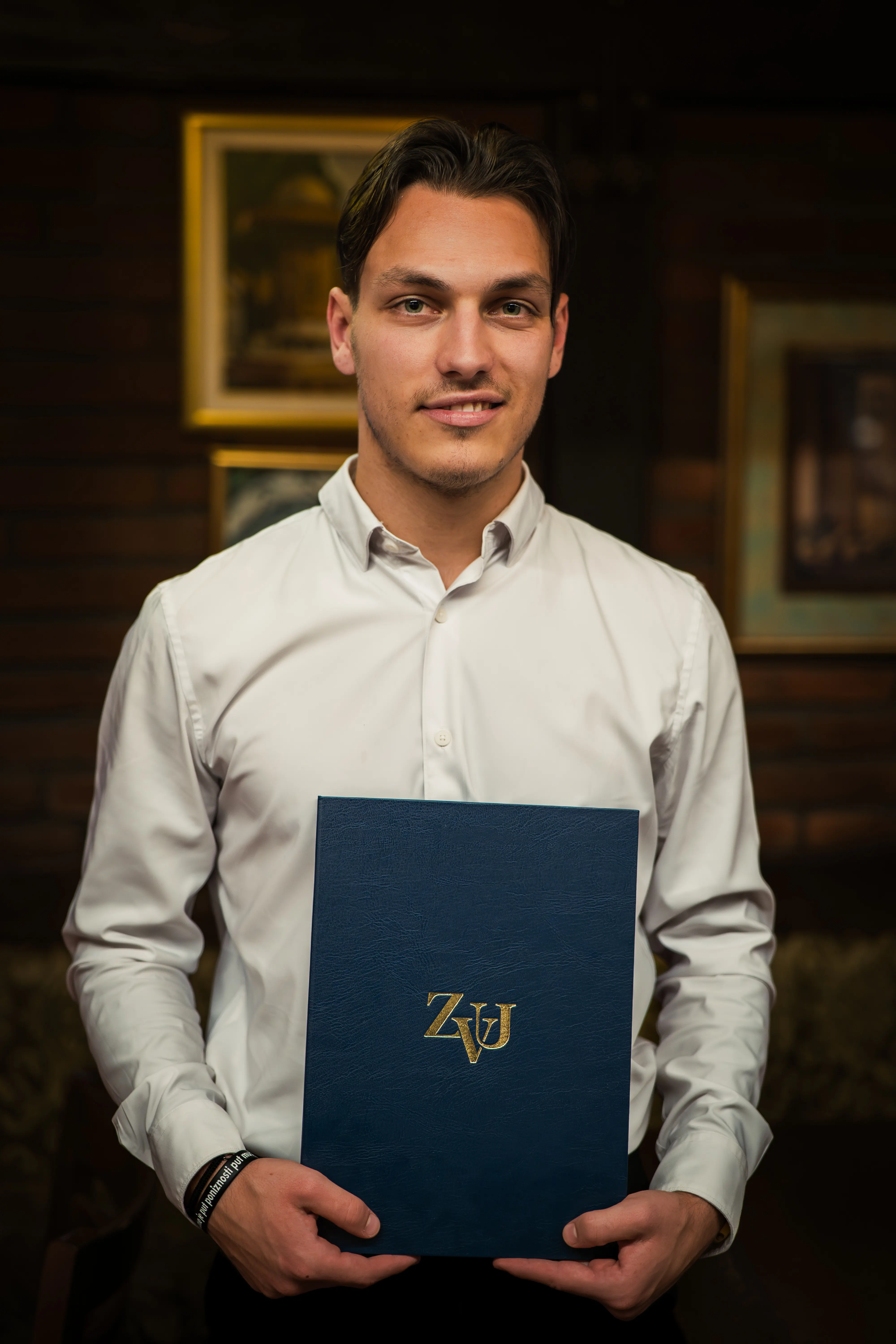 Formal portrait of a graduate presenting a blue certificate folder against softly lit interior decor.
